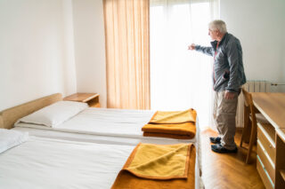 photo - Elderly Man in Hotel Room Looking out Window