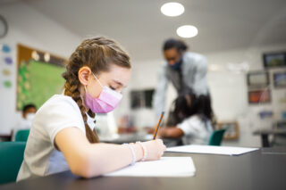 photo - Elementary Student Writing with Pencil at Desk with Teacher in Background