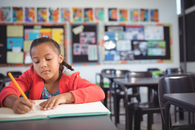 photo - Girl Writing While Sitting at Desk in Classroom